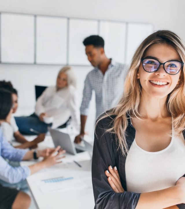 blonde female executive posing with smile and arms crossed during brainstorm with managers indoor portrait of european student spending time in hall with asian and african friends min scaled 1